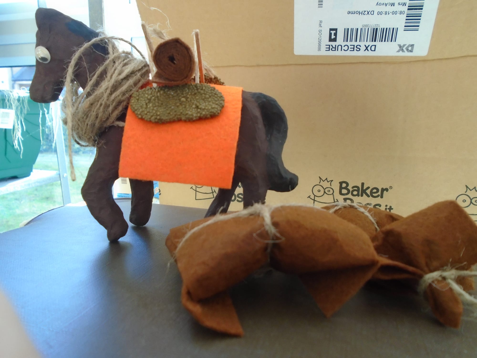 Wobbly-eyed model pack horse with orange felt saddle blanket, panniers, and a coil of rope round its neck.