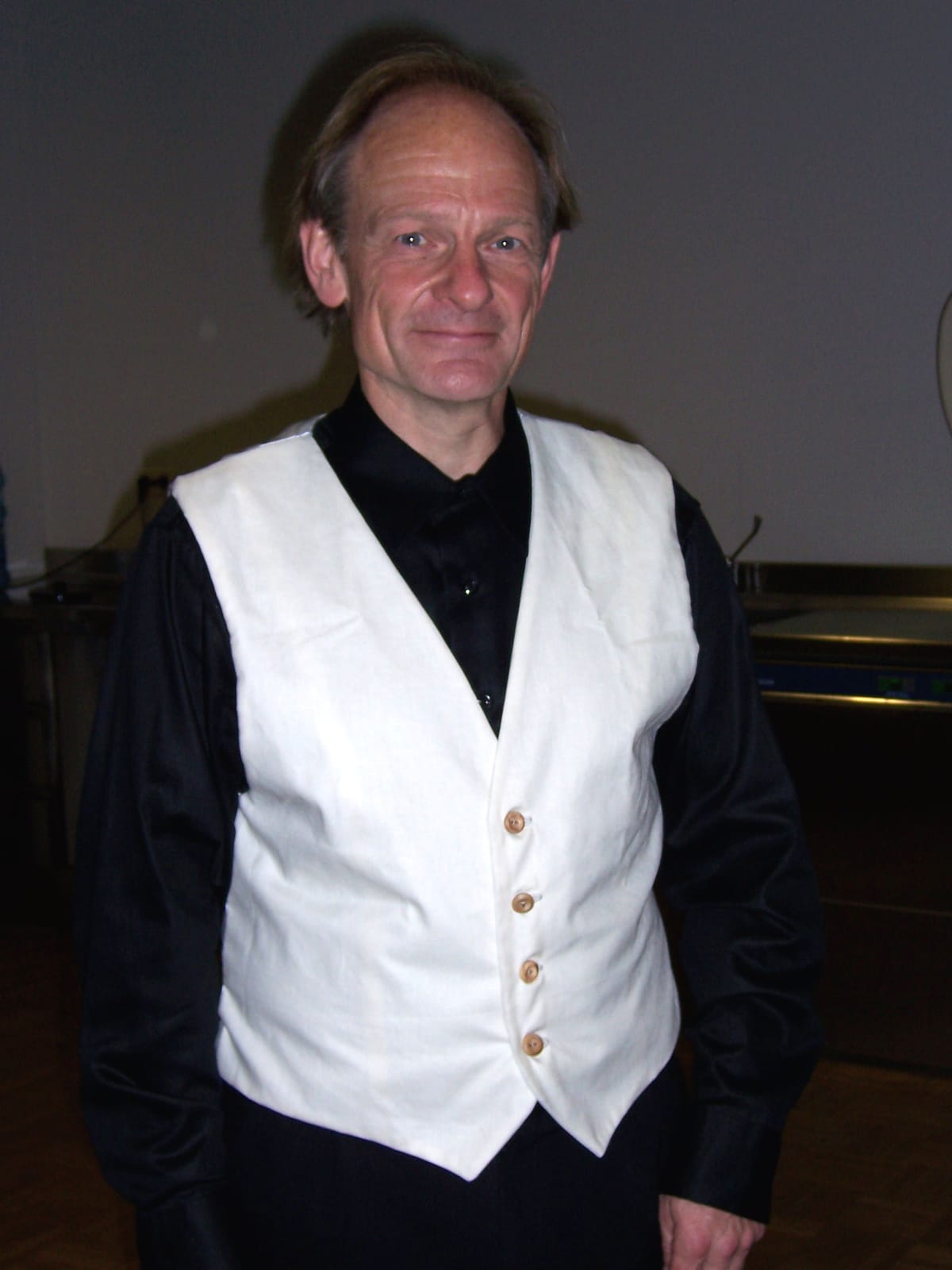 A rather bald gentleman wearing an off-white calico waistcoat over black everything else.