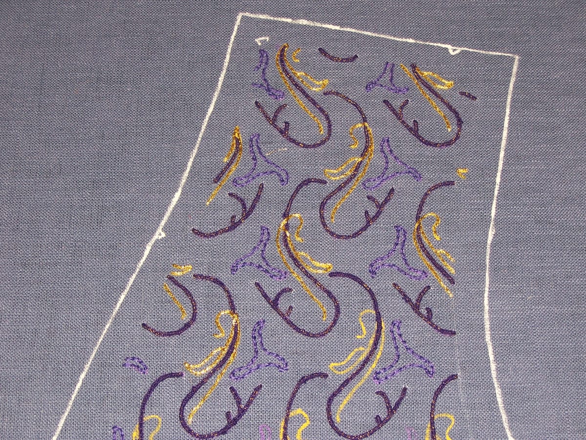 Purple and gold embroidery on air-force blue fabric. The design is regularly repeating and somewhat botanical.