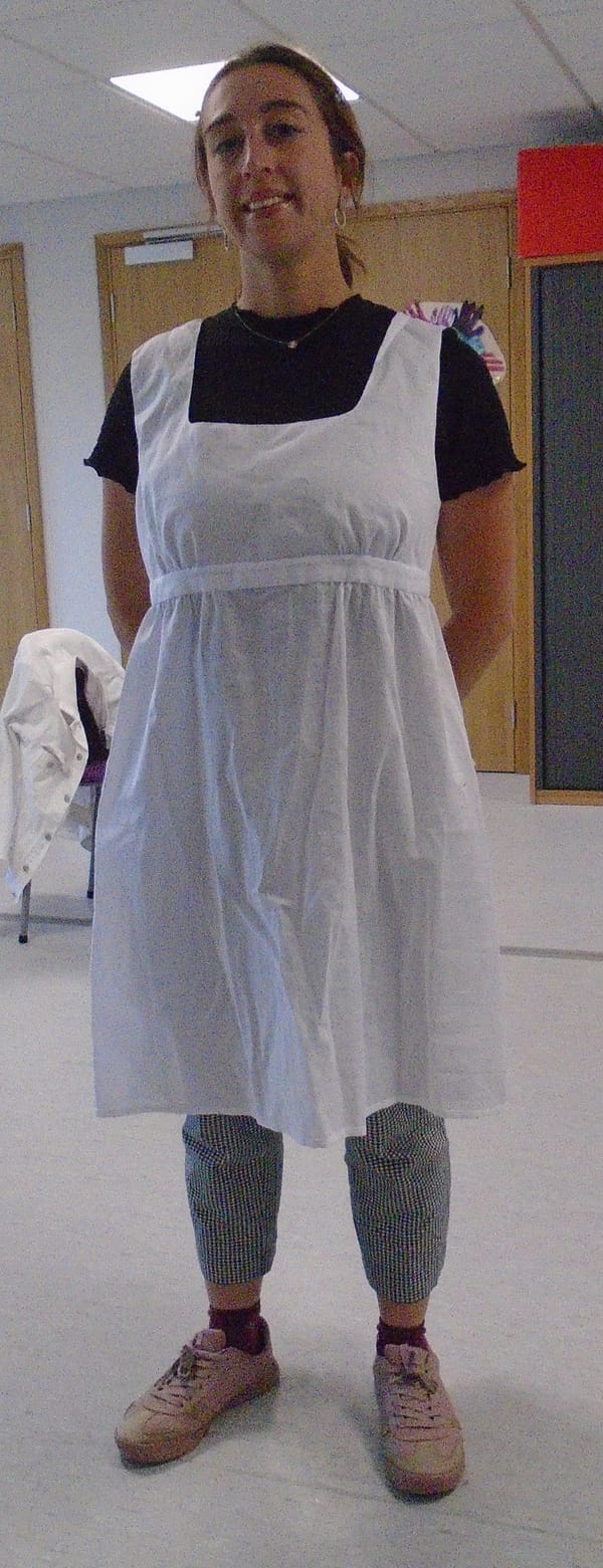 Alice wears the pinafore over a black T-shirt, cropped trousers with a tiny check, and brown lace-up shoes.