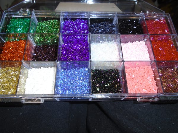 18-compartment organiser containing a selection of bugle beads.  Most of the compartments are quite full.