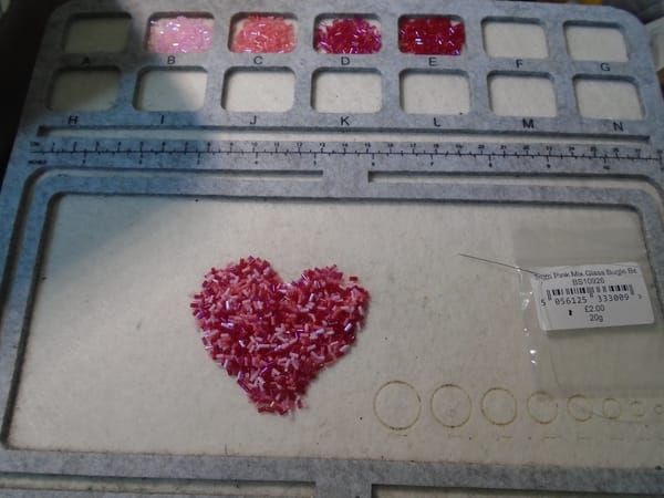 A pile of mixed pink 3 mm bugle beads, shaped into a heart.  Above can be seen the four constituent shades.