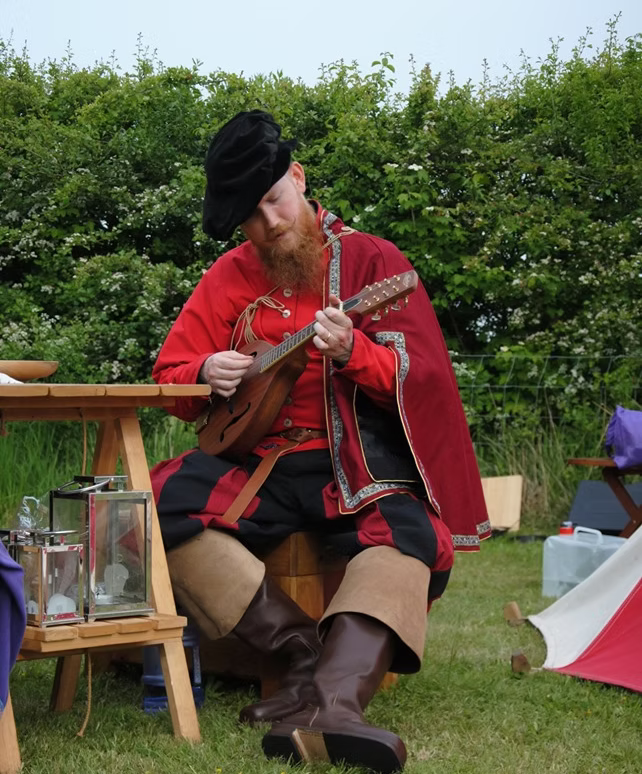 SCA bard with mandolin (?); he is dressed mostly in red, with a black hat and brown foldover boots.