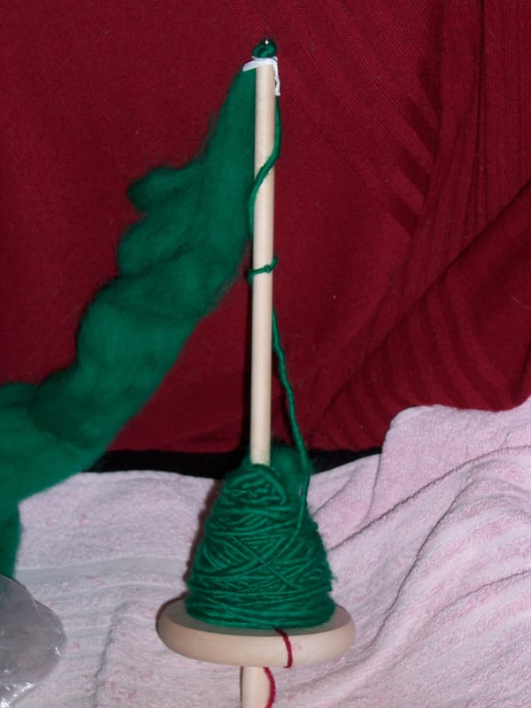 About 50 g of green yarn wound onto a drop spindle.  There is also a piece of red yarn at the bottom (the "leader").